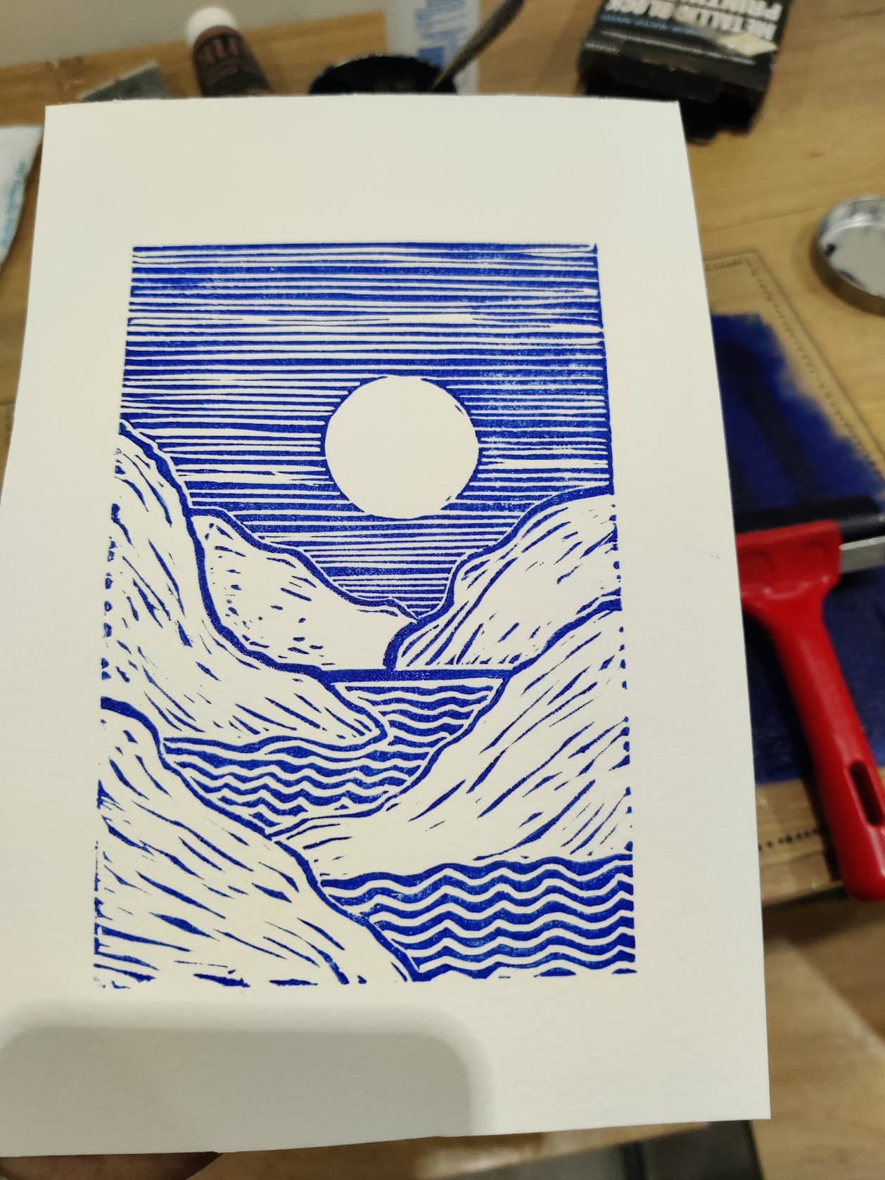 A blue lino print of a sun over a mountain valley and sea, held up next to a red brayer