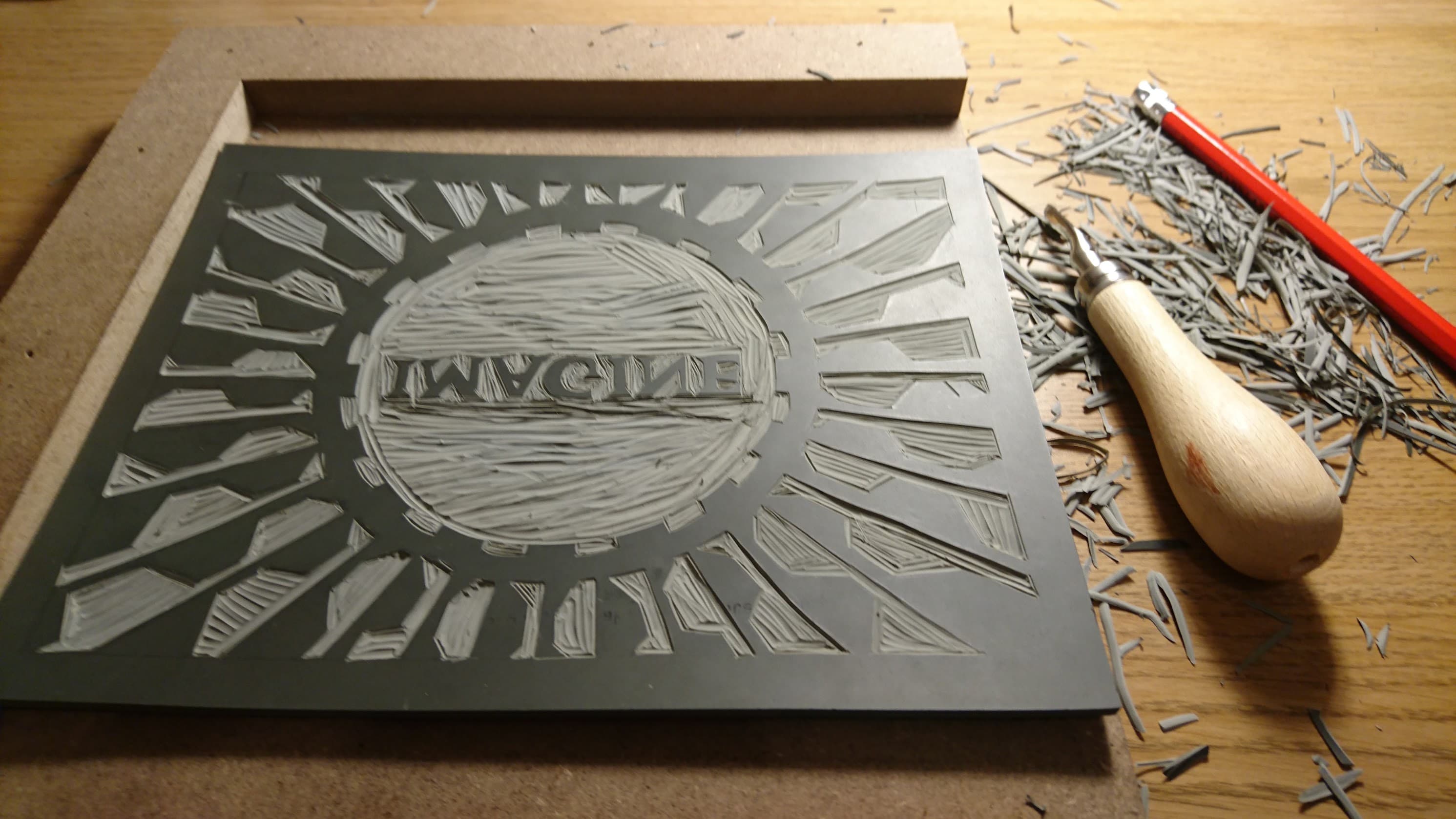 A carved lino block with the word Imagine cut backwards in the middle of a sunburst, next to a carving tool and rubber shavings