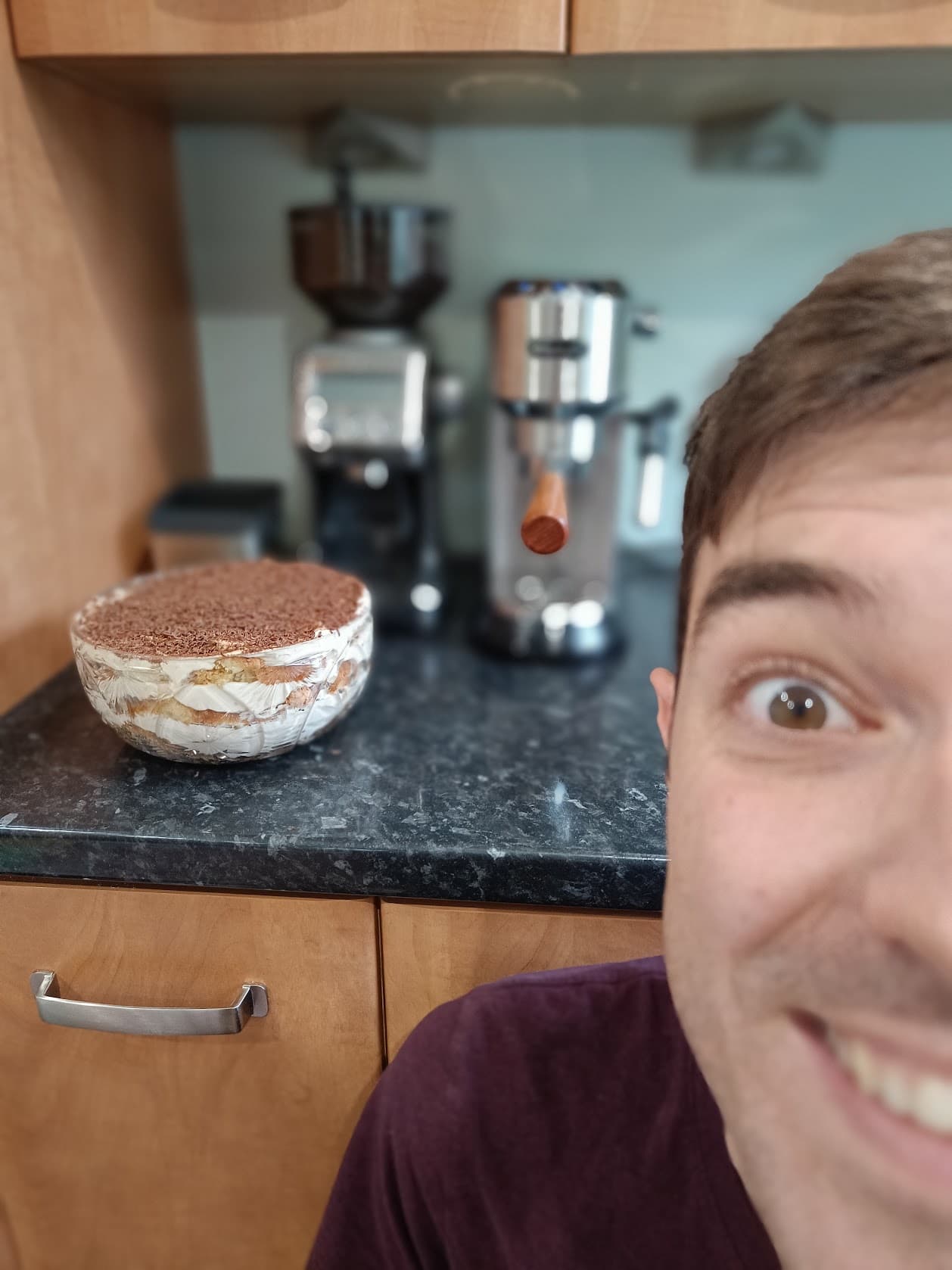 Jamie grinning at the camera next to a homemade tiramisu, a coffee grinder and an espresso machine
