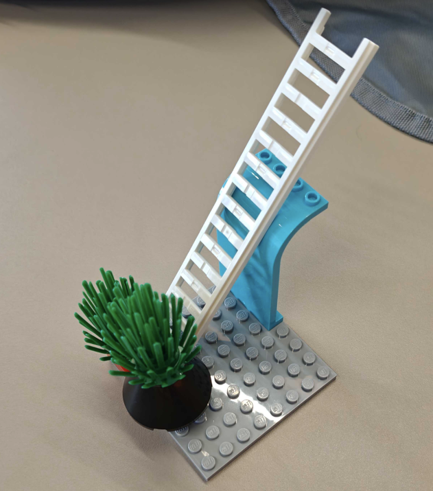 A small Lego model: a grey baseplate with a green plant in a pot, and a white ladder leaning on a blue curved brick